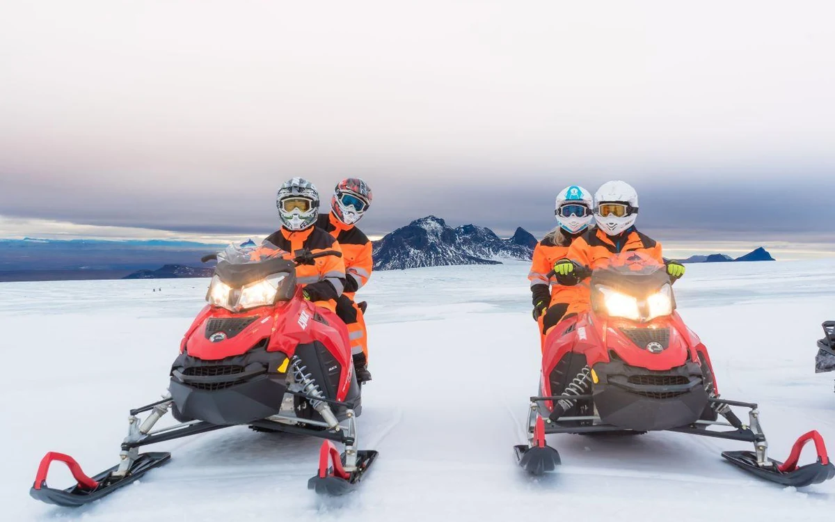 Dos motos de nieve sobre nieve plana, cada una con dos ocupantes con monos naranjas